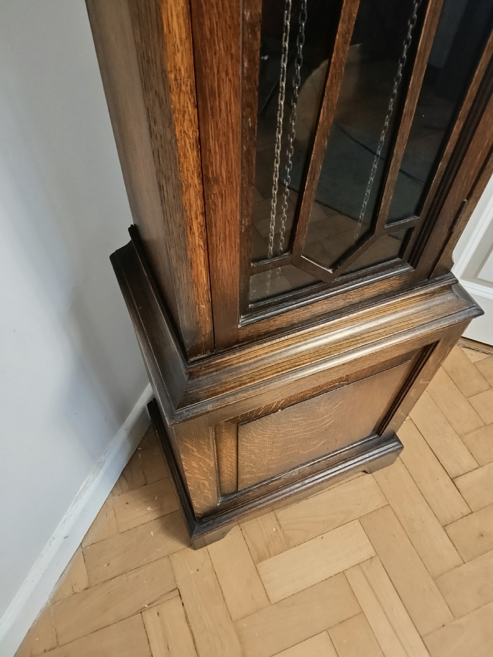 A tall dark oak longcase clock featuring an arched bonnet, barley twist side columns, a brass dial with Roman numerals, and a glazed trunk door displaying three brass weights and pendulum. View VI