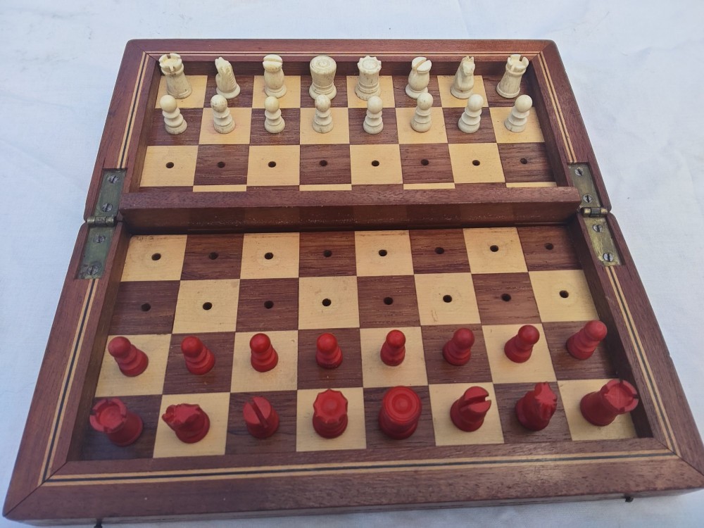 An open antique mahogany folding travelling chess board containing miniature carved red and natural white bone chess pieces fitted into individual pegged holes.