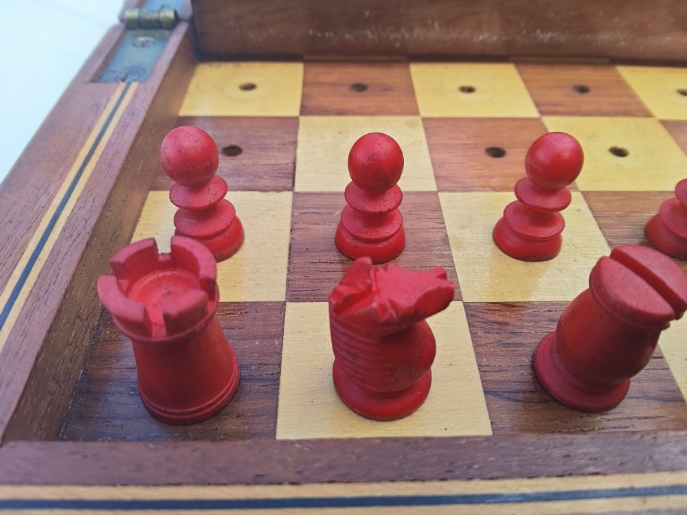 An open antique mahogany folding travelling chess board containing miniature carved red and natural white bone chess pieces fitted into individual pegged holes. View IX