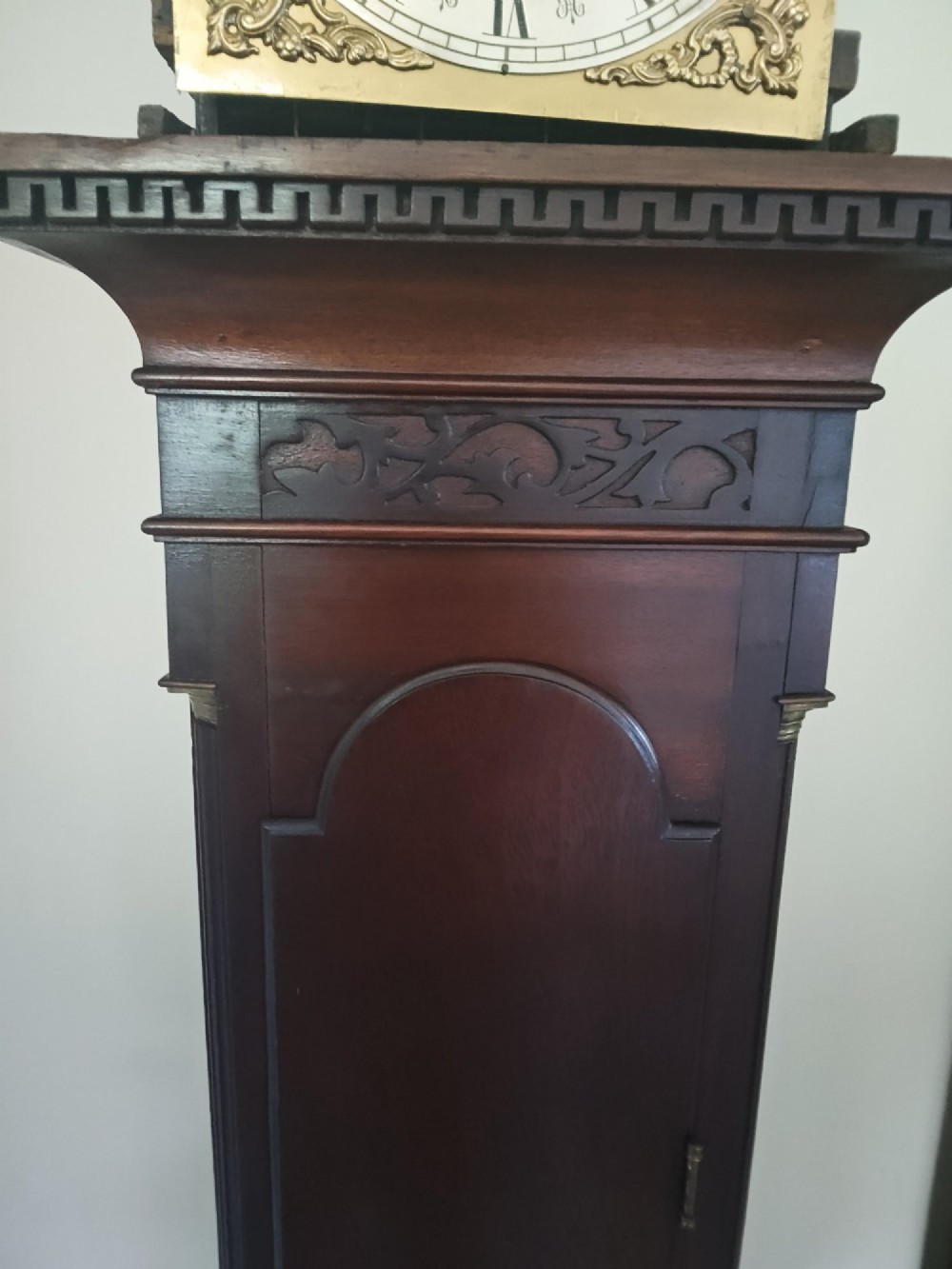 A slim dark mahogany longcase clock featuring a swan-neck pediment, brass dial with Roman numerals, and an arched trunk door. View III