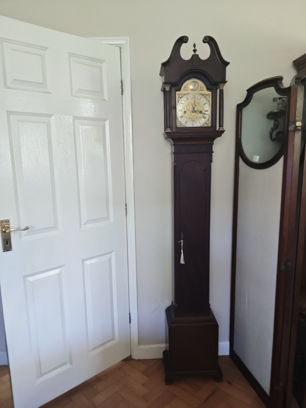 A slim dark mahogany longcase clock featuring a swan-neck pediment, brass dial with Roman numerals, and an arched trunk door. View XI
