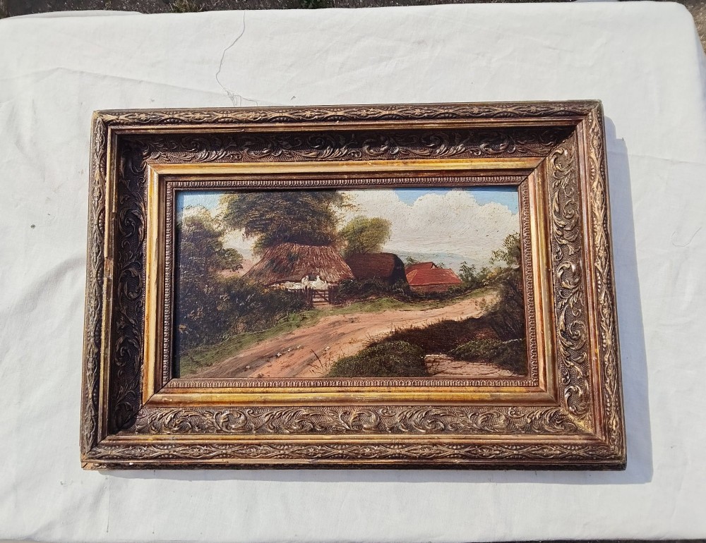 An antique oil painting on card depicting a rural English thatched cottage along a dirt path, set inside an ornate gilt frame.