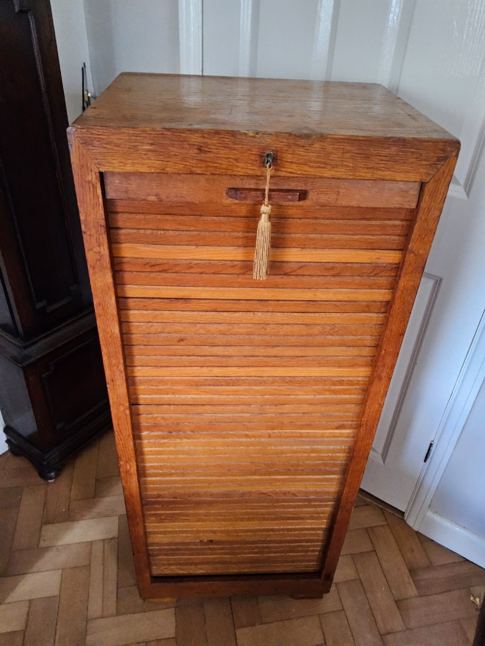 A tall oak tambour filing cabinet featuring a slatted wooden roll-front shutter door with a key inserted near the upper handle. View II