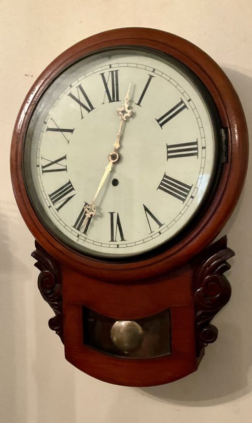 A 19th-century American mahogany drop dial wall clock featuring a circular dial with Roman numerals, carved side scrolls, and a lower glass aperture displaying a metal pendulum.