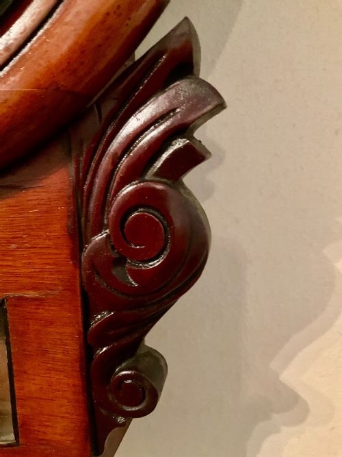 A 19th-century American mahogany drop dial wall clock featuring a circular dial with Roman numerals, carved side scrolls, and a lower glass aperture displaying a metal pendulum. View II