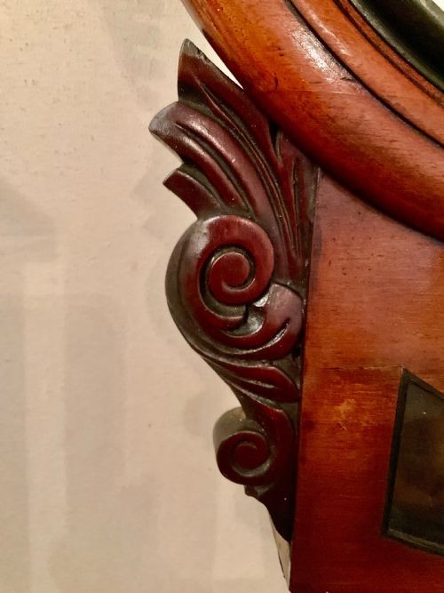 A 19th-century American mahogany drop dial wall clock featuring a circular dial with Roman numerals, carved side scrolls, and a lower glass aperture displaying a metal pendulum. View III