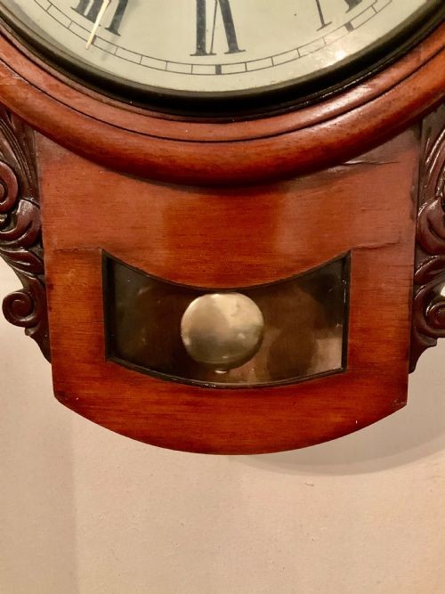 A 19th-century American mahogany drop dial wall clock featuring a circular dial with Roman numerals, carved side scrolls, and a lower glass aperture displaying a metal pendulum. View IV