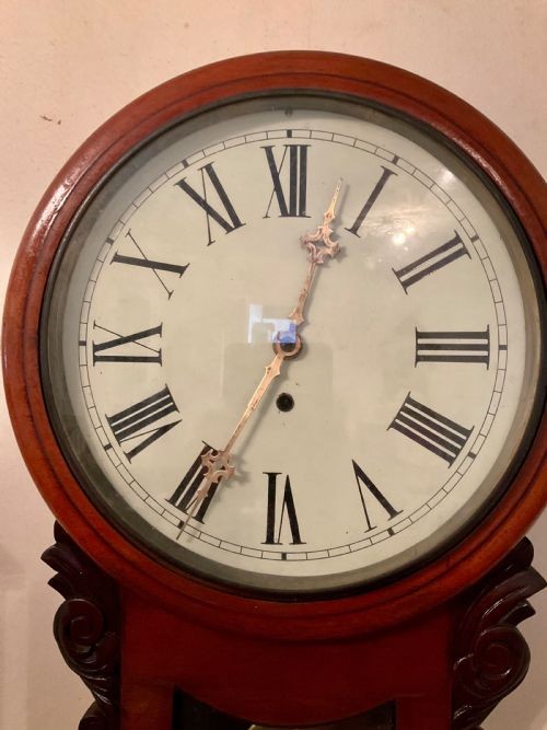 A 19th-century American mahogany drop dial wall clock featuring a circular dial with Roman numerals, carved side scrolls, and a lower glass aperture displaying a metal pendulum. View V