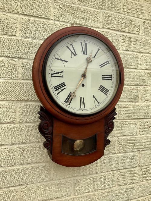 A 19th-century American mahogany drop dial wall clock featuring a circular dial with Roman numerals, carved side scrolls, and a lower glass aperture displaying a metal pendulum. View VI