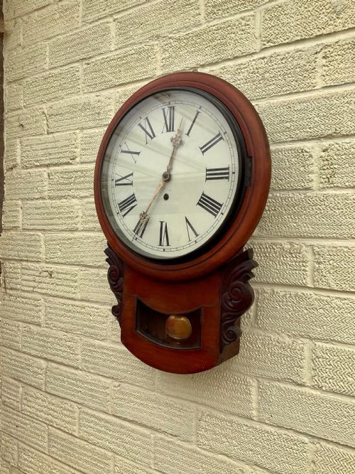 A 19th-century American mahogany drop dial wall clock featuring a circular dial with Roman numerals, carved side scrolls, and a lower glass aperture displaying a metal pendulum. View VIII