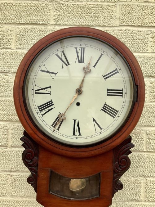 A 19th-century American mahogany drop dial wall clock featuring a circular dial with Roman numerals, carved side scrolls, and a lower glass aperture displaying a metal pendulum. View IX