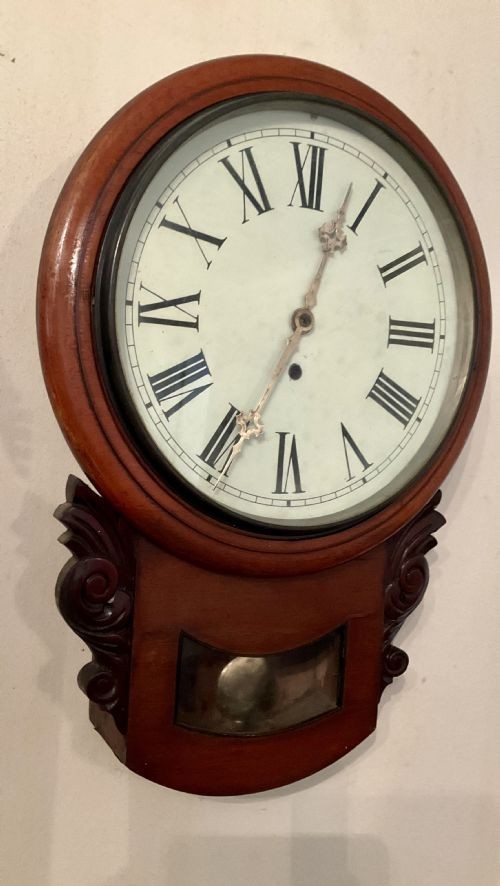 A 19th-century American mahogany drop dial wall clock featuring a circular dial with Roman numerals, carved side scrolls, and a lower glass aperture displaying a metal pendulum. View X