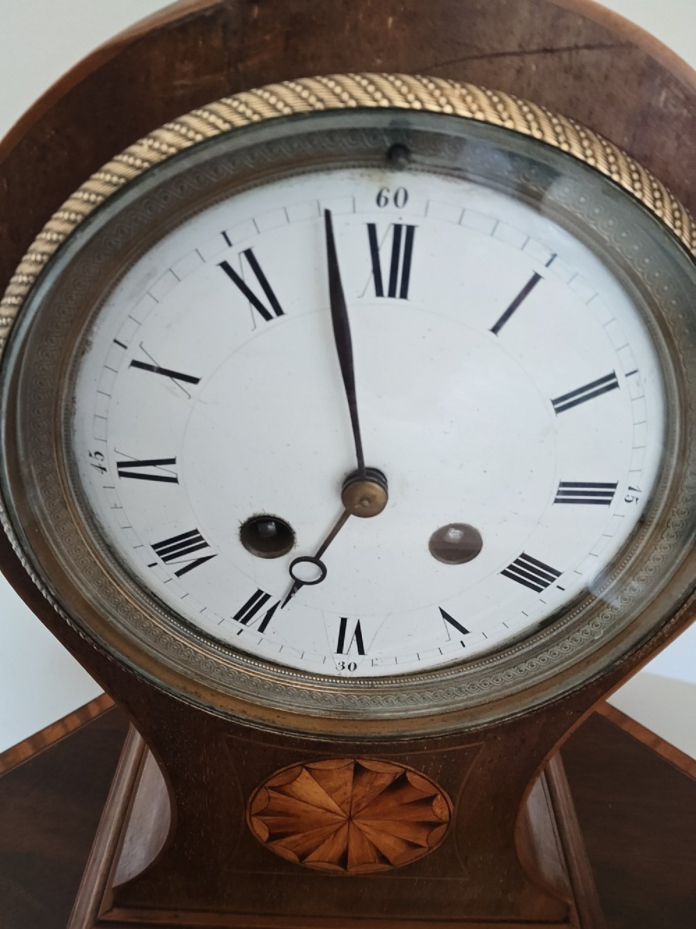 An antique balloon-shaped mahogany mantel clock featuring a white enamel dial with Roman numerals, wood marquetry inlay, and small brass feet. View VI