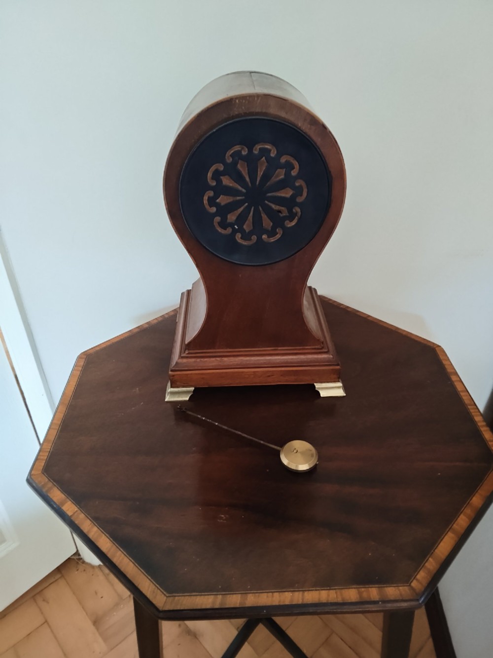 An antique balloon-shaped mahogany mantel clock featuring a white enamel dial with Roman numerals, wood marquetry inlay, and small brass feet. View IX