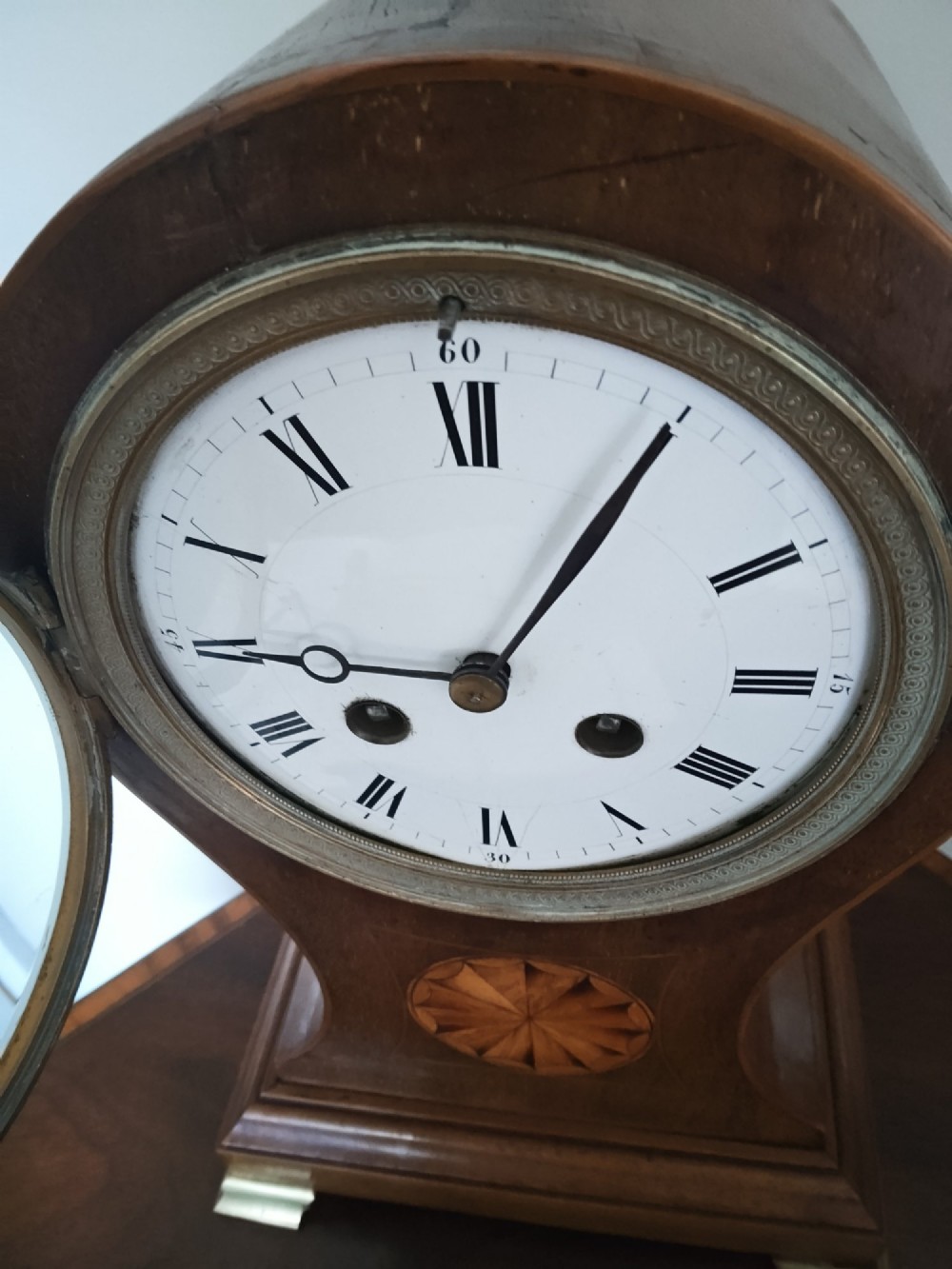 An antique balloon-shaped mahogany mantel clock featuring a white enamel dial with Roman numerals, wood marquetry inlay, and small brass feet. View X