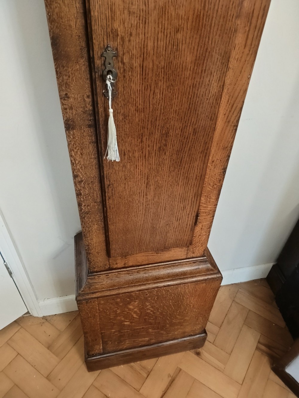 A freestanding Georgian oak longcase clock featuring a square brass dial, slender trunk door, turned hood columns, and a square plinth base. View V