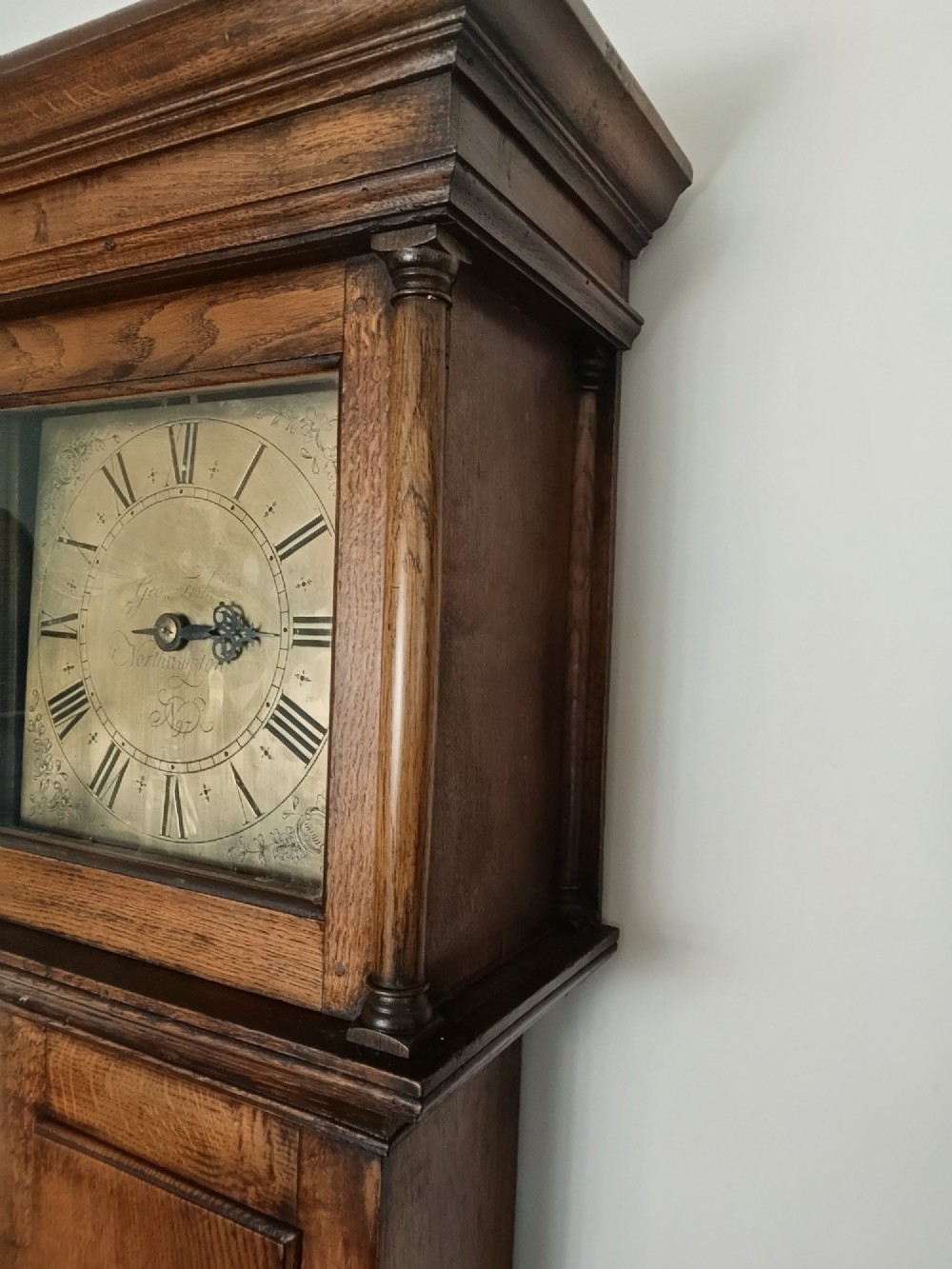 A freestanding Georgian oak longcase clock featuring a square brass dial, slender trunk door, turned hood columns, and a square plinth base. View XI