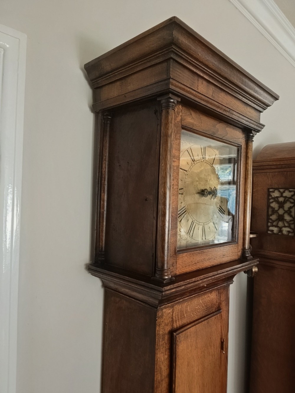 A freestanding Georgian oak longcase clock featuring a square brass dial, slender trunk door, turned hood columns, and a square plinth base. View XIII