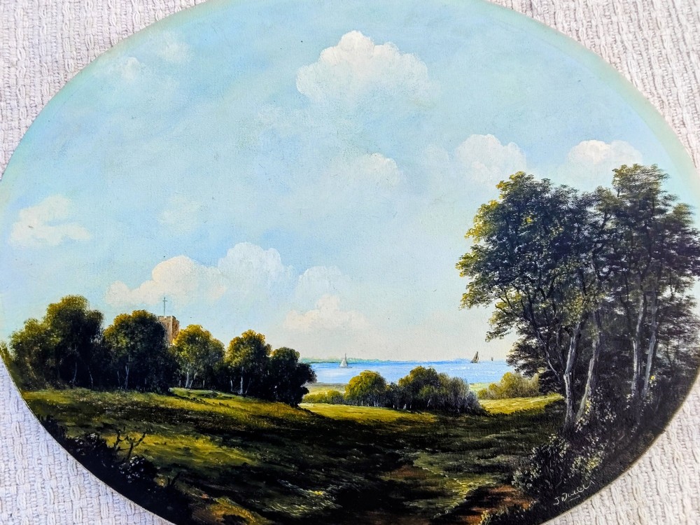 An oval oil painting depicting a grassy coastal landscape with trees, a distant church tower, and sailboats on the blue sea beneath a cloudy sky, encased in a gilt frame. View IV