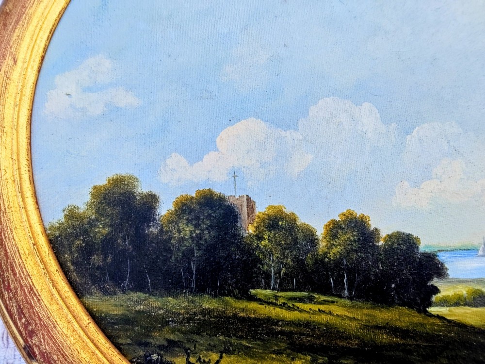 An oval oil painting depicting a grassy coastal landscape with trees, a distant church tower, and sailboats on the blue sea beneath a cloudy sky, encased in a gilt frame. View V