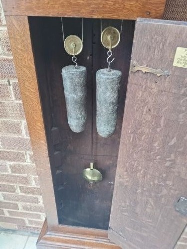 An upright wooden longcase clock featuring a square brass dial, glass door hood, and a narrow oak trunk standing against a textured brick wall. View XI