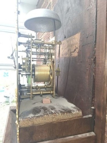 An upright wooden longcase clock featuring a square brass dial, glass door hood, and a narrow oak trunk standing against a textured brick wall. View II