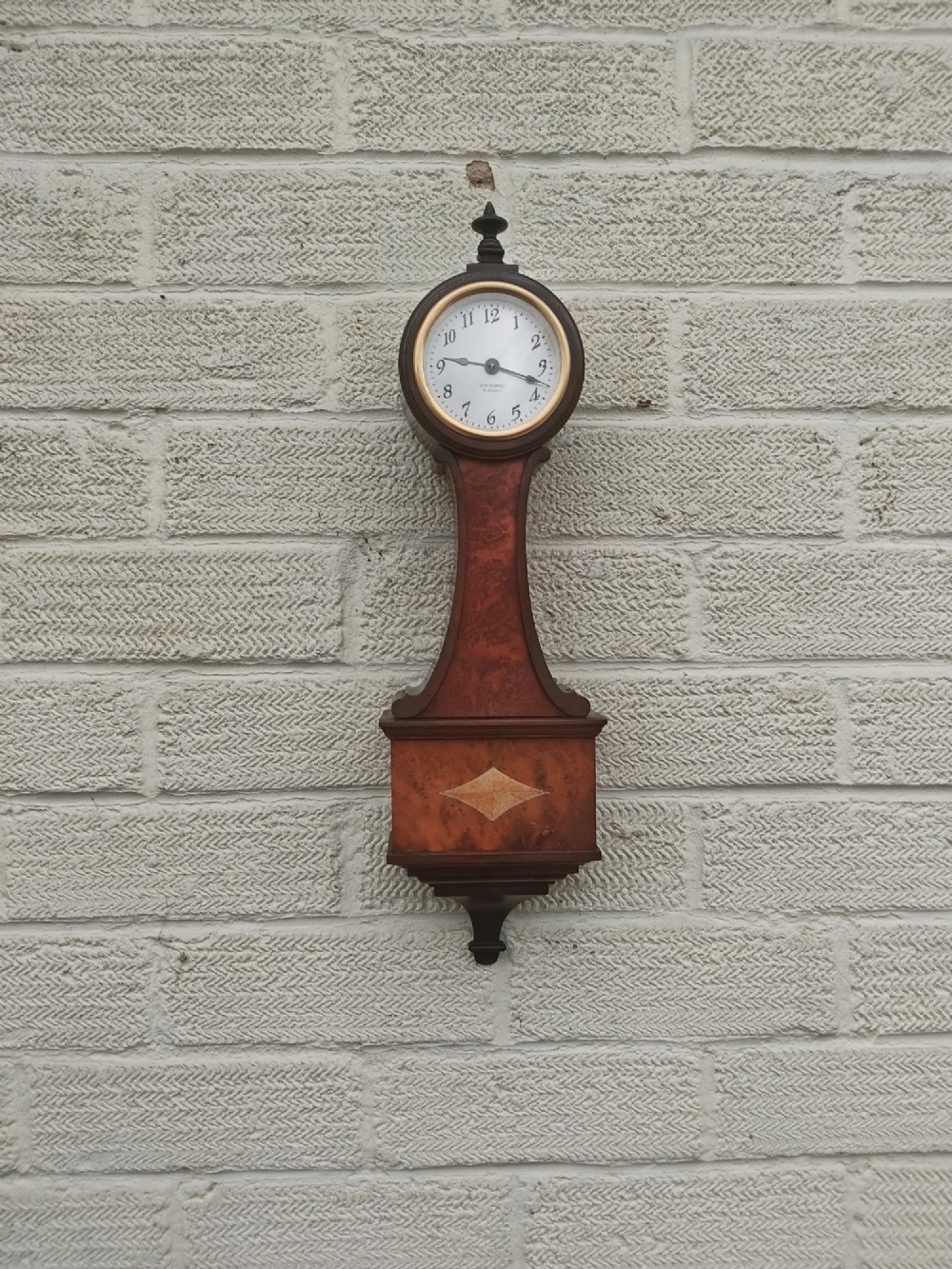 A vertical wooden banjo wall clock featuring a round white dial with black numerals, a narrow waist, and a rectangular lower section with a diamond inlay.