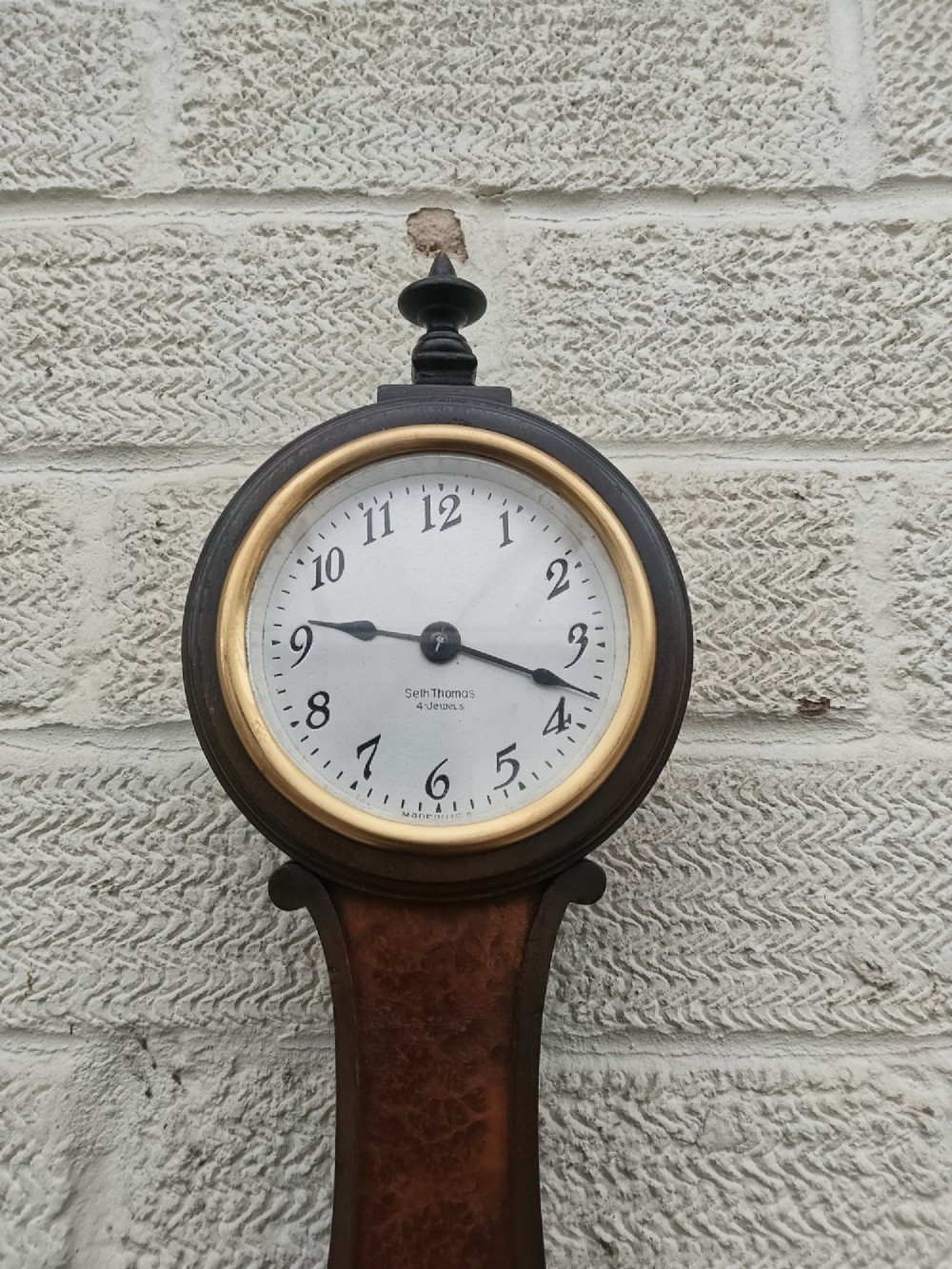 A vertical wooden banjo wall clock featuring a round white dial with black numerals, a narrow waist, and a rectangular lower section with a diamond inlay. View V