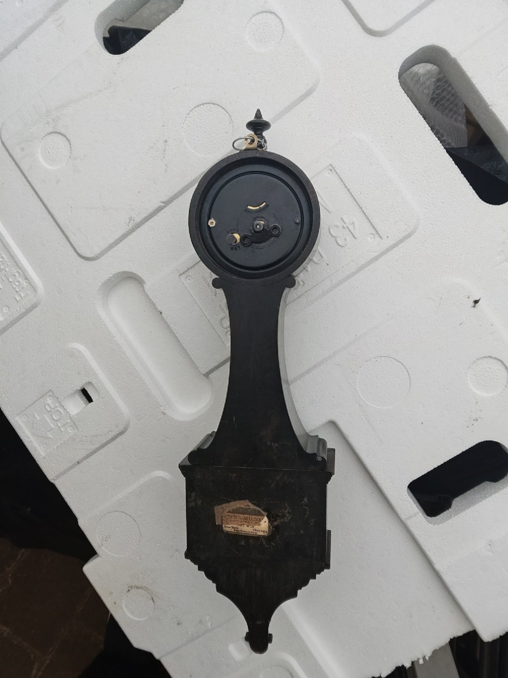 A vertical wooden banjo wall clock featuring a round white dial with black numerals, a narrow waist, and a rectangular lower section with a diamond inlay. View X