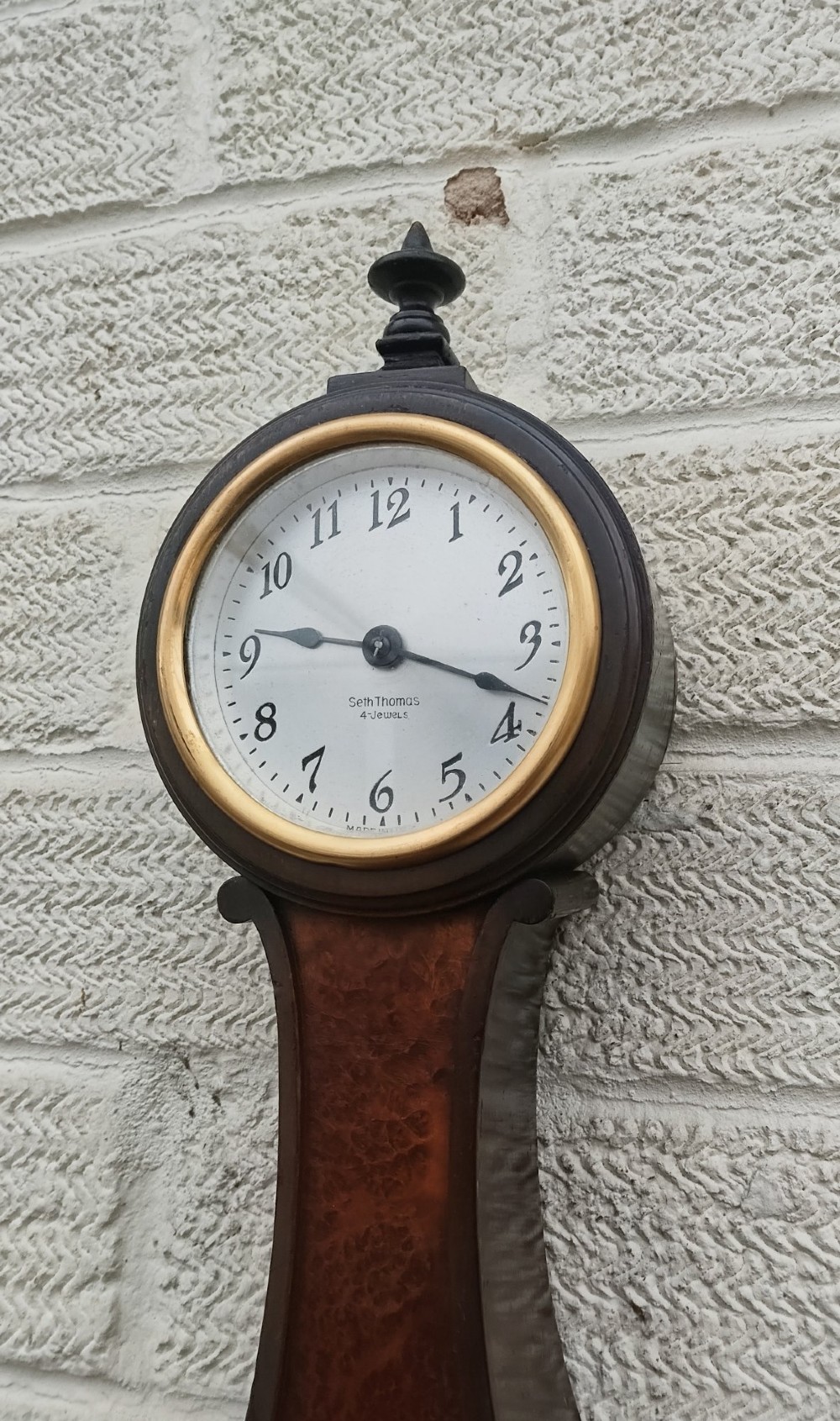 A vertical wooden banjo wall clock featuring a round white dial with black numerals, a narrow waist, and a rectangular lower section with a diamond inlay. View XI