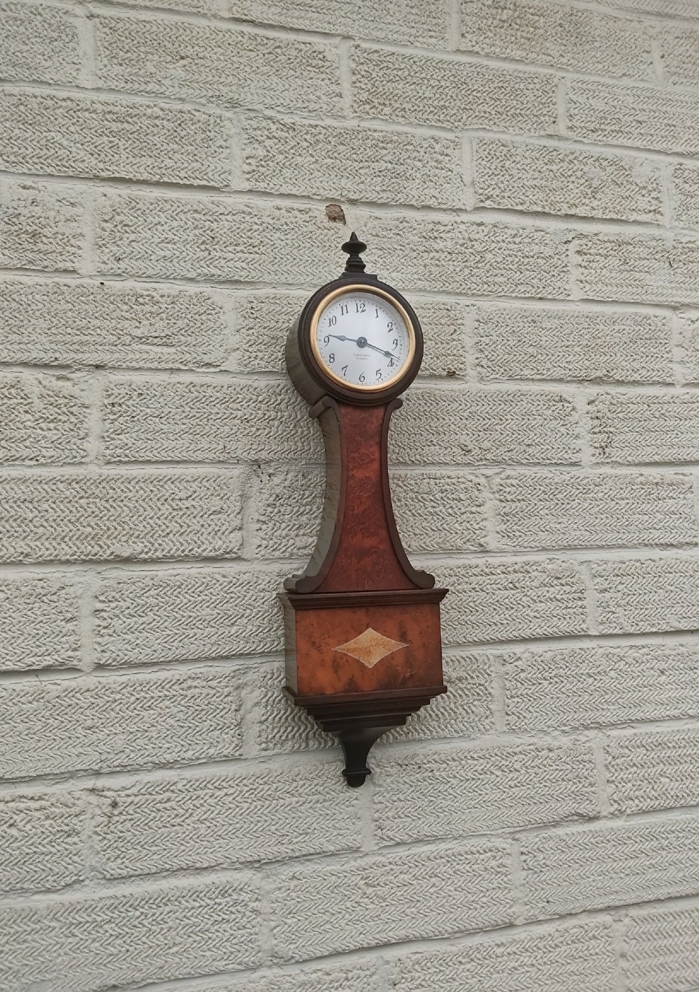 A vertical wooden banjo wall clock featuring a round white dial with black numerals, a narrow waist, and a rectangular lower section with a diamond inlay. View XII