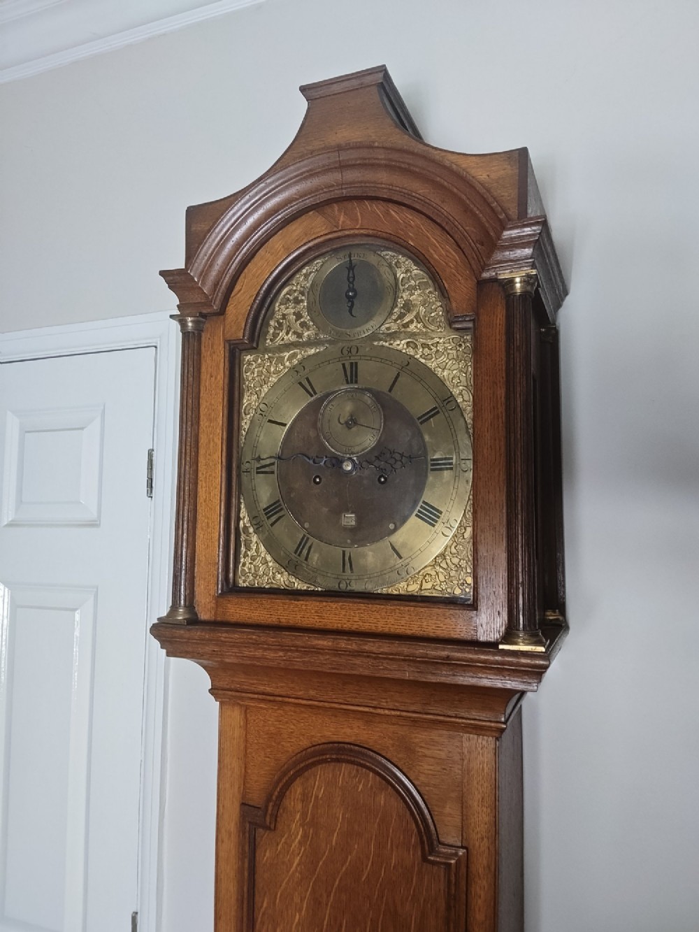 An Early Georgian floor-standing oak longcase clock featuring a carved arch hood, brass dial with strike and silent selector, and a slim trunk door. View III