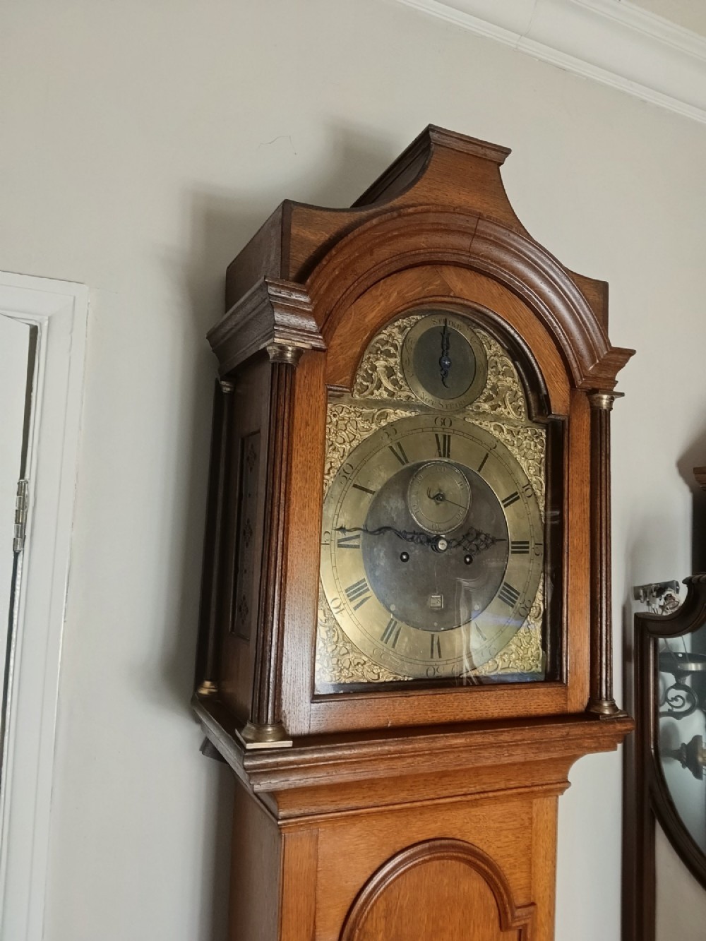 An Early Georgian floor-standing oak longcase clock featuring a carved arch hood, brass dial with strike and silent selector, and a slim trunk door. View IV