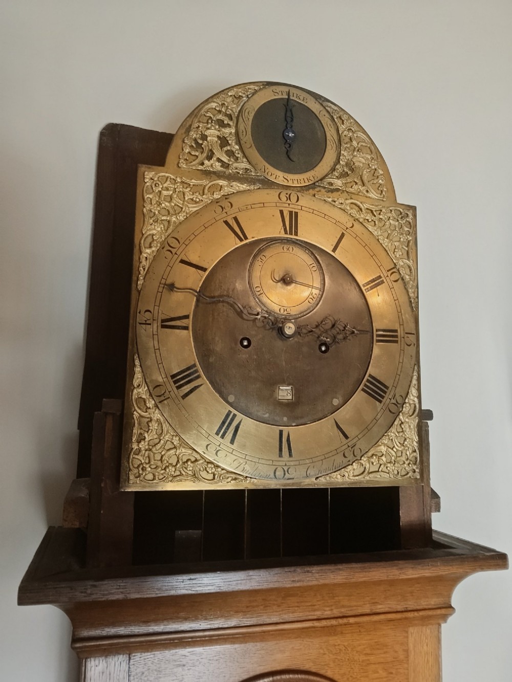 An Early Georgian floor-standing oak longcase clock featuring a carved arch hood, brass dial with strike and silent selector, and a slim trunk door. View VII