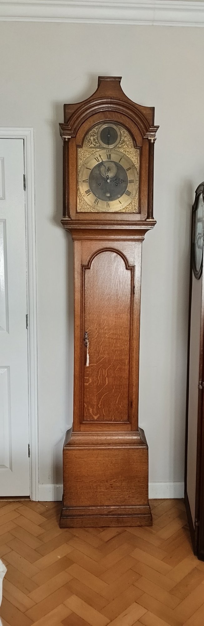 An Early Georgian floor-standing oak longcase clock featuring a carved arch hood, brass dial with strike and silent selector, and a slim trunk door. View X