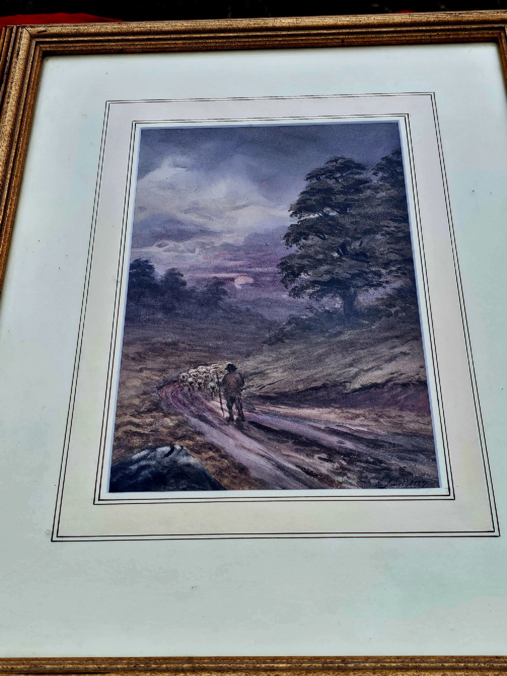 A framed watercolour painting depicting a shepherd driving a flock of sheep along a dirt track under a purple twilight sky.