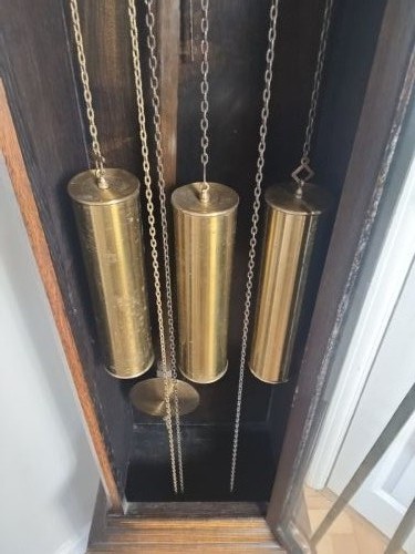 A tall dark oak longcase clock featuring an arched bonnet, barley twist side columns, a brass dial with Roman numerals, and a glazed trunk door displaying three brass weights and pendulum. View VII