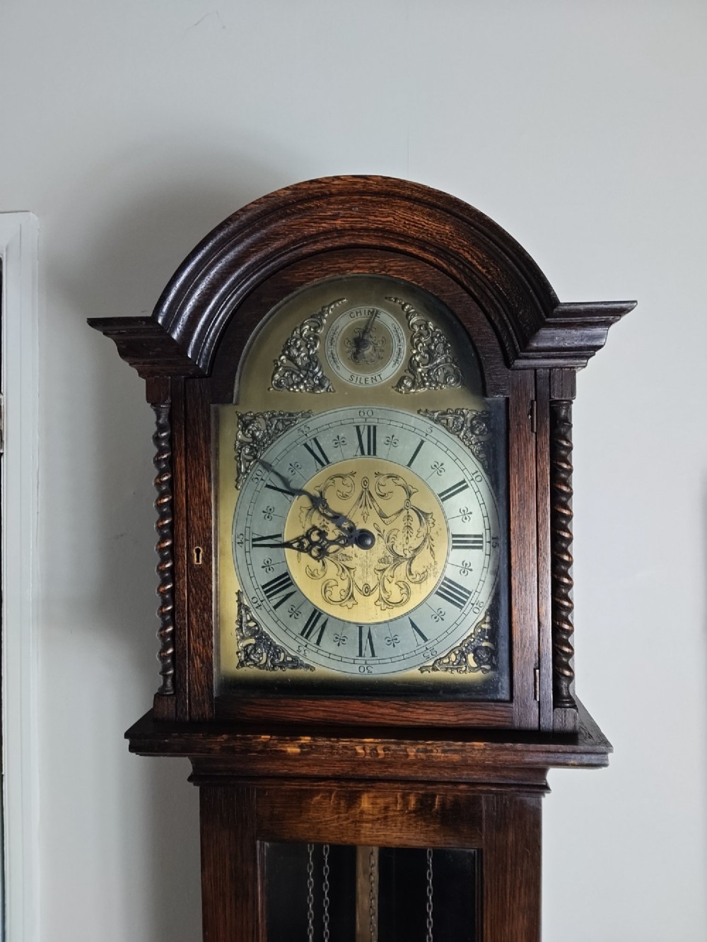 A 1920s dark oak grandmother longcase clock featuring an arched top hood, barley twist columns, brass dial, and glazed door displaying weights. View II