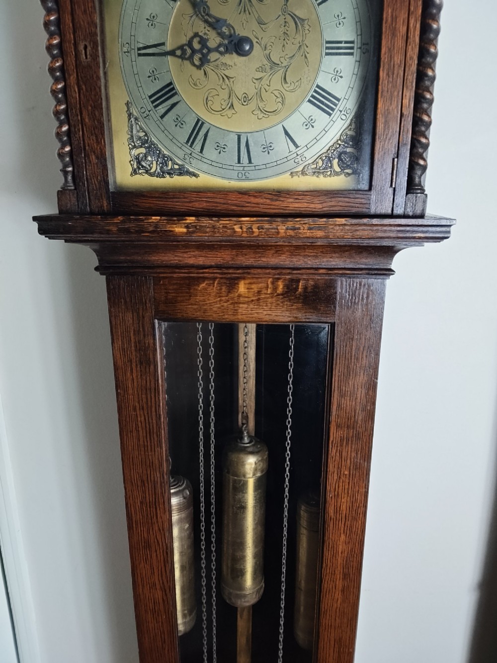 A 1920s dark oak grandmother longcase clock featuring an arched top hood, barley twist columns, brass dial, and glazed door displaying weights. View IV