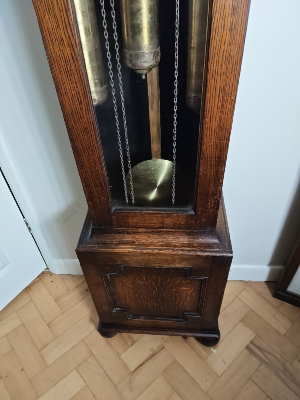A 1920s dark oak grandmother longcase clock featuring an arched top hood, barley twist columns, brass dial, and glazed door displaying weights. View V