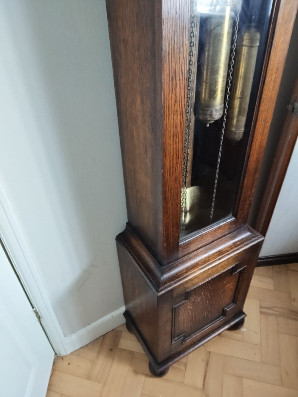 A 1920s dark oak grandmother longcase clock featuring an arched top hood, barley twist columns, brass dial, and glazed door displaying weights. View X