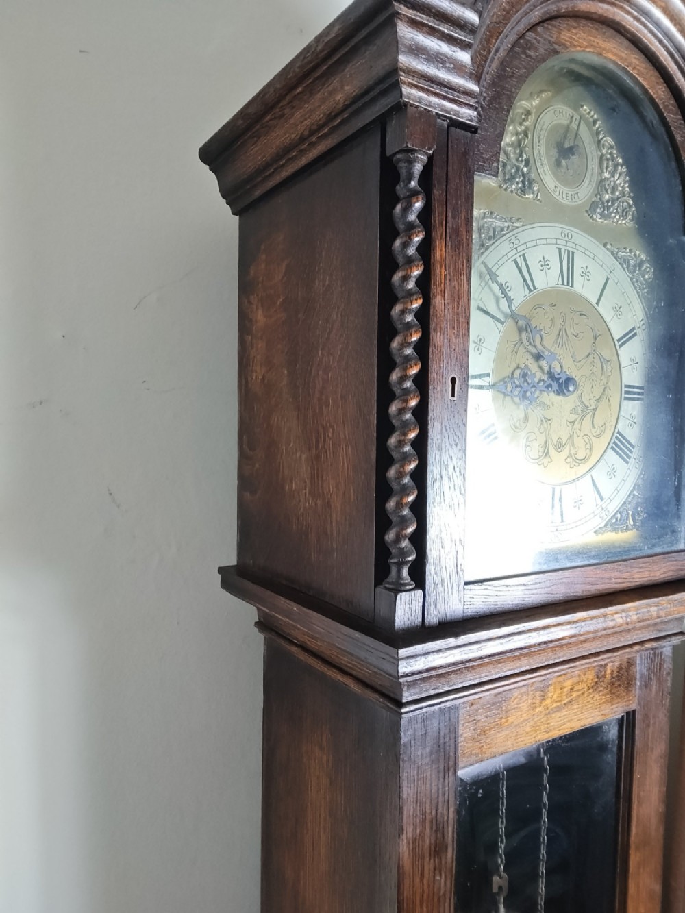 A 1920s dark oak grandmother longcase clock featuring an arched top hood, barley twist columns, brass dial, and glazed door displaying weights. View XI