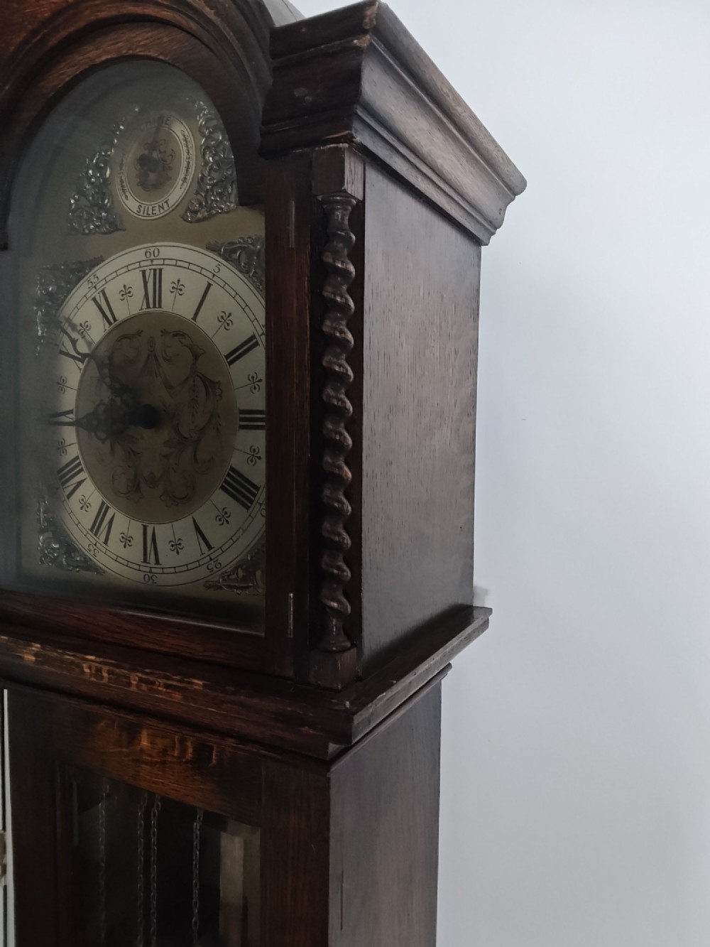 A 1920s dark oak grandmother longcase clock featuring an arched top hood, barley twist columns, brass dial, and glazed door displaying weights. View XII