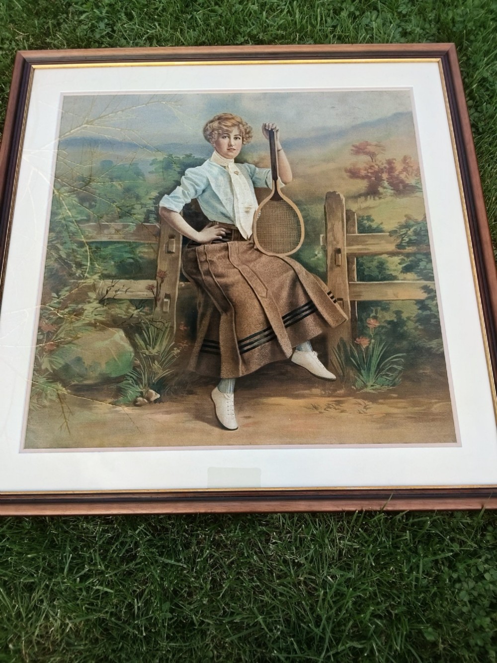 A framed Edwardian tapestry depicting a woman in early twentieth-century sporting attire holding a tennis racket beside a wooden fence. View II