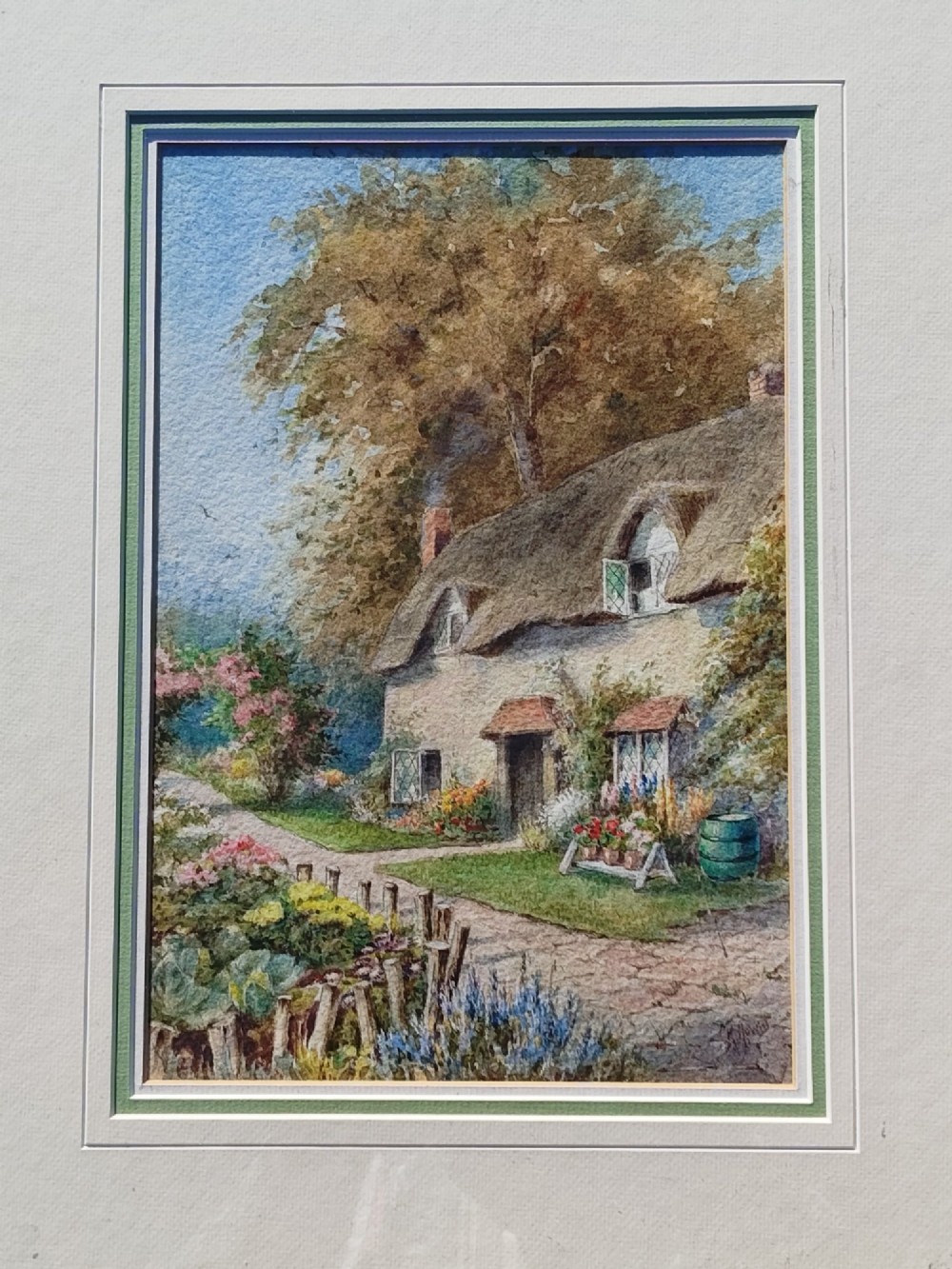 A framed vertical watercolour painting depicting a traditional thatched cottage with a garden filled with colourful flowers under a blue sky.