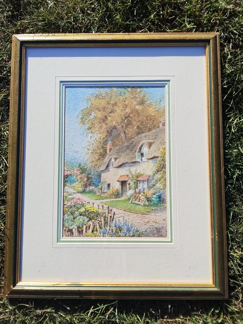 A framed vertical watercolour painting depicting a traditional thatched cottage with a garden filled with colourful flowers under a blue sky. View II