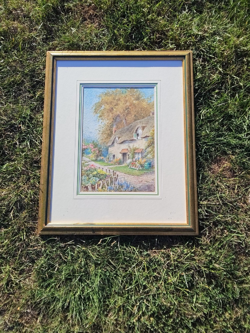 A framed vertical watercolour painting depicting a traditional thatched cottage with a garden filled with colourful flowers under a blue sky. View III