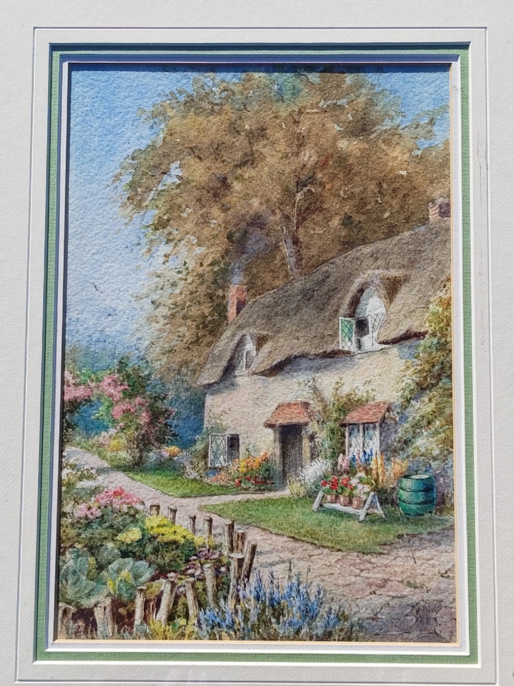 A framed vertical watercolour painting depicting a traditional thatched cottage with a garden filled with colourful flowers under a blue sky. View VI