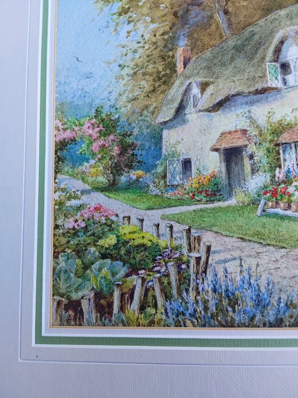 A framed vertical watercolour painting depicting a traditional thatched cottage with a garden filled with colourful flowers under a blue sky. View X