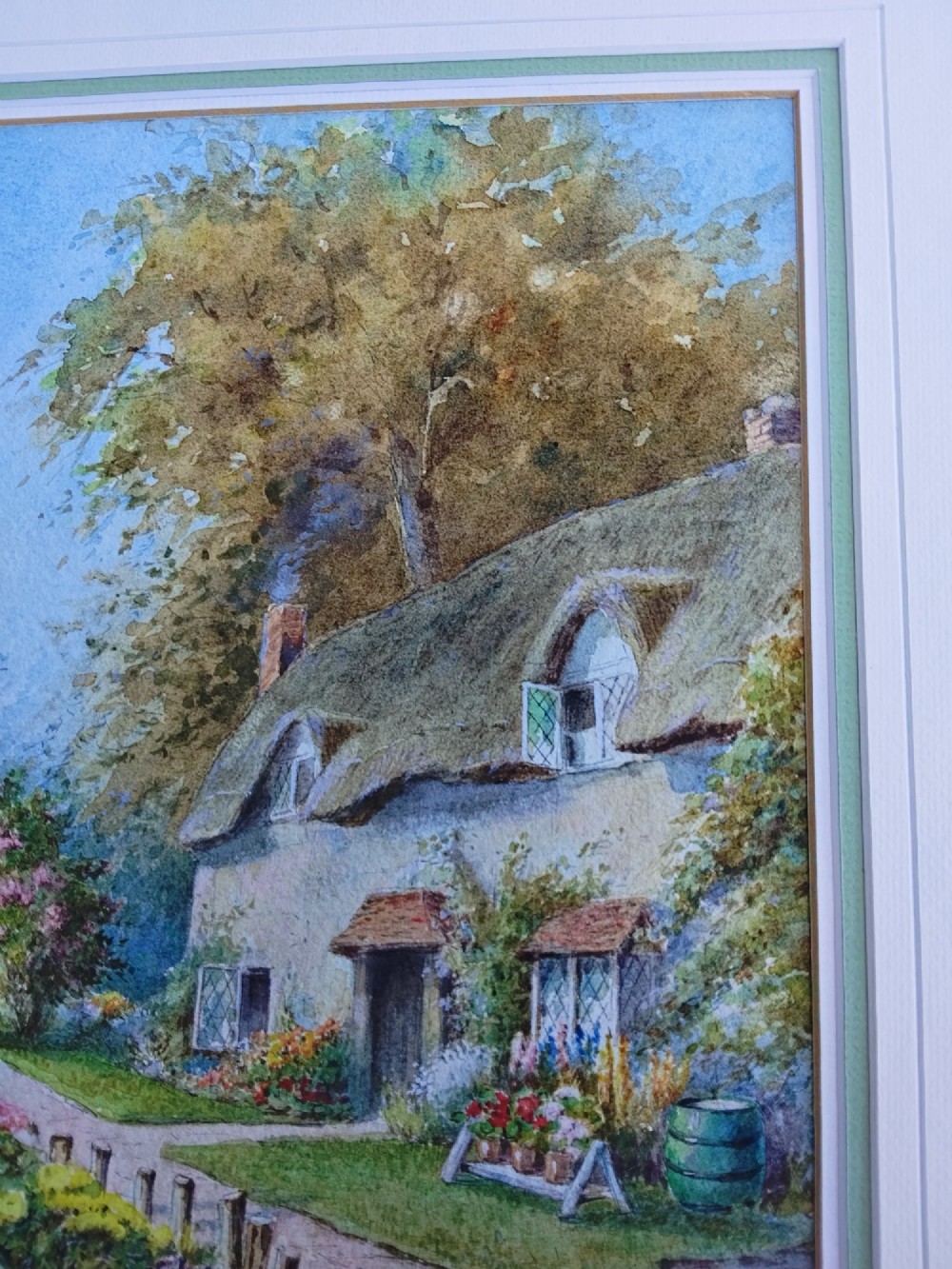 A framed vertical watercolour painting depicting a traditional thatched cottage with a garden filled with colourful flowers under a blue sky. View XI